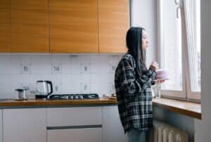 A woman standing alone in her kitchen, thoughtfully gazing out the window.