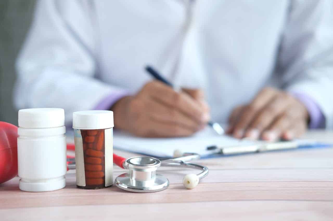 Close-up of doctor writing prescription with medicines and stethoscope on desk