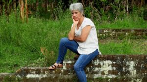 An older woman in casual attire sits outdoors, looking sad